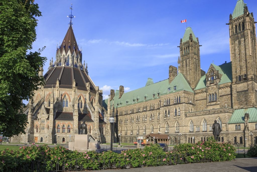 Historic Parliament Buildings with stone towers and green roofs under a blue sky, seen from a garden.