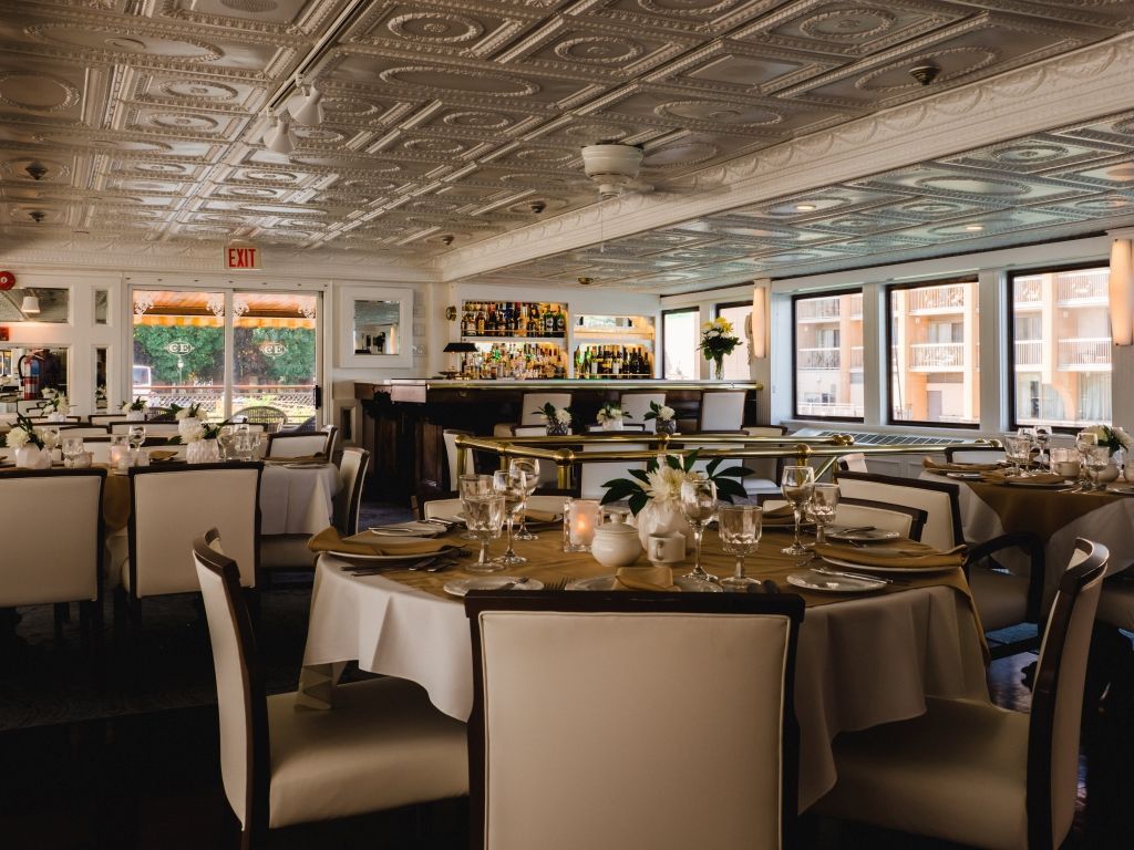 Elegant restaurant dining room with set tables, white chairs, ornate ceiling, and large windows