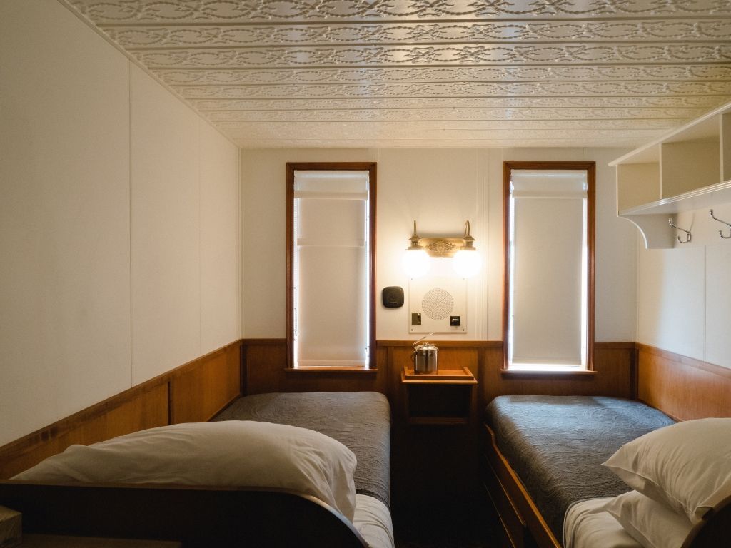 Small cozy bedroom with two single beds, wood paneling, and two windows by a desk