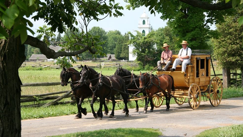 Horse-drawn carriage on a tree-lined path, with two passengers and a driver passing a white church