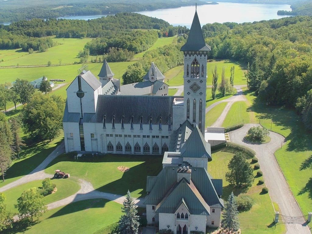 An aerial view of a tall stone church with a steeple surrounded by green fields, trees, and a lake.