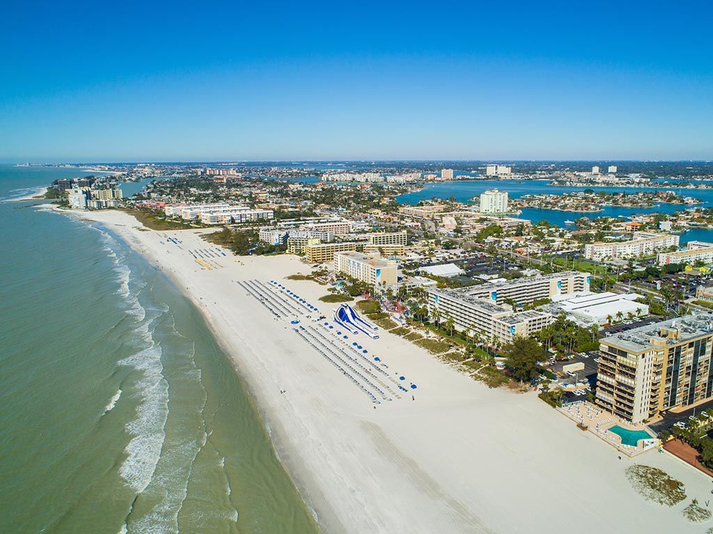 Aerial view of a white sandy beach with turquoise water and a city skyline under a blue sky.