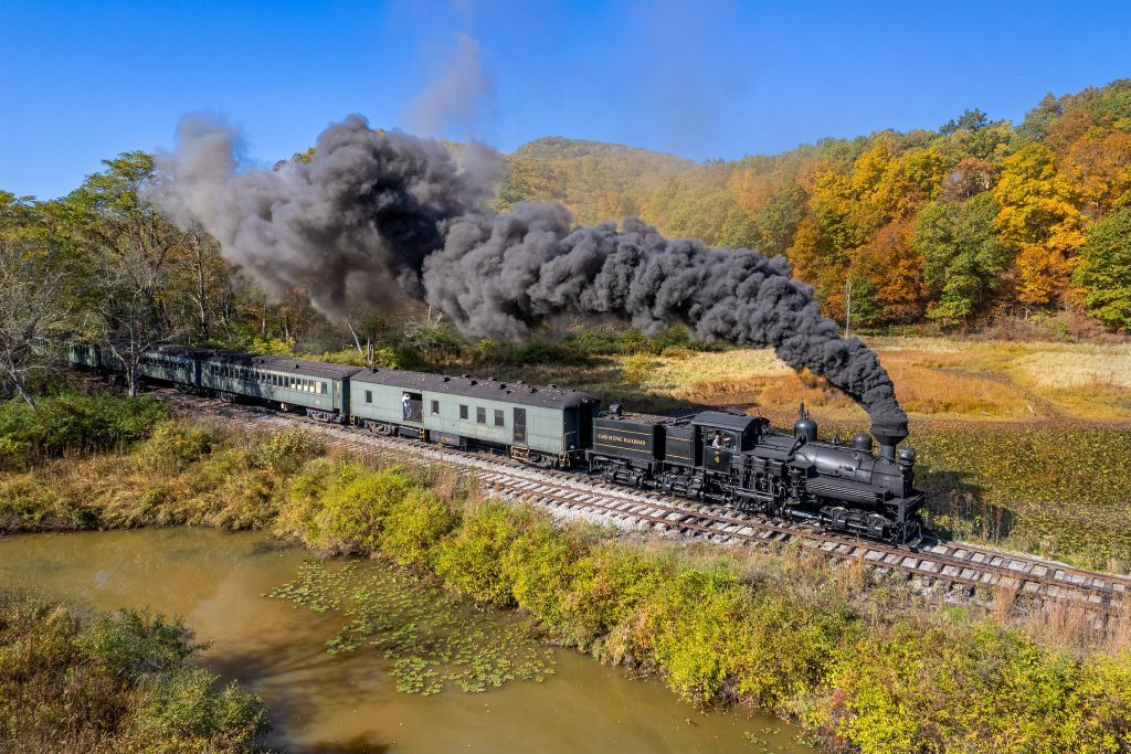 Steam train billowing black smoke, traveling through a colorful autumn landscape over a water way.