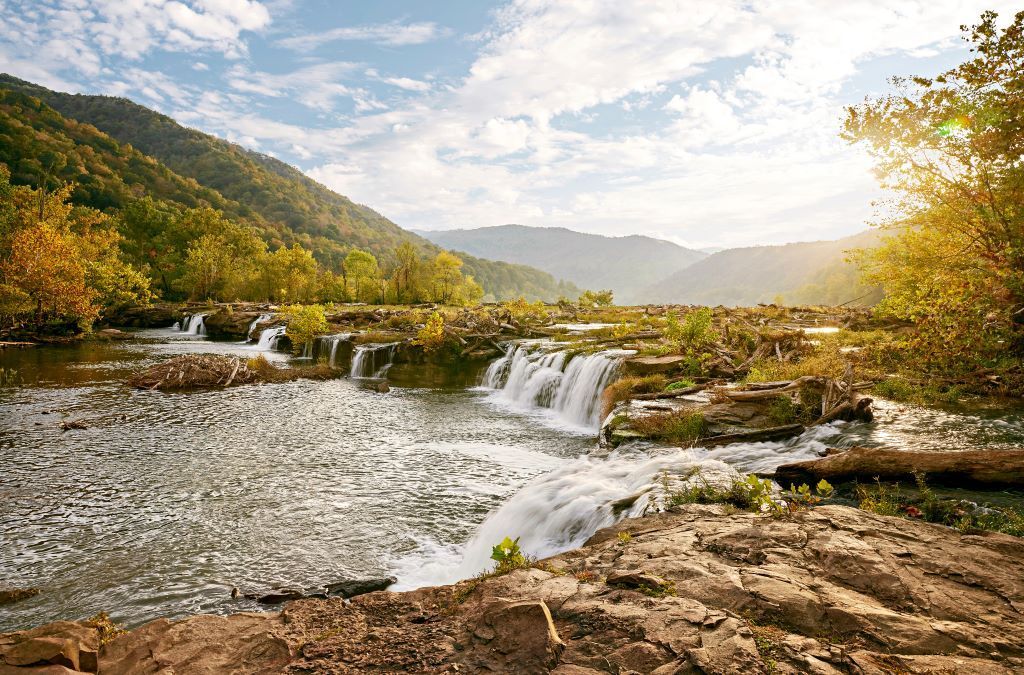 A river cascades over rocks, framed by mountains and trees under a bright, sunny sky.