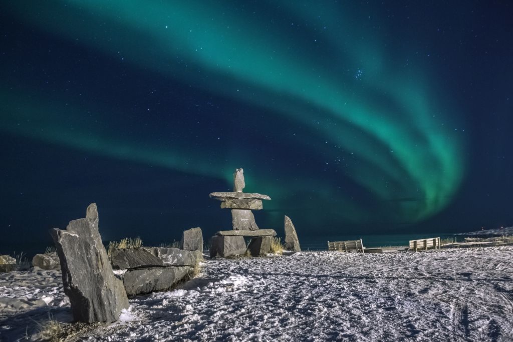 Aurora borealis over an Inukshuk on a snow-covered landscape. Green and blue lights against a starry night sky.