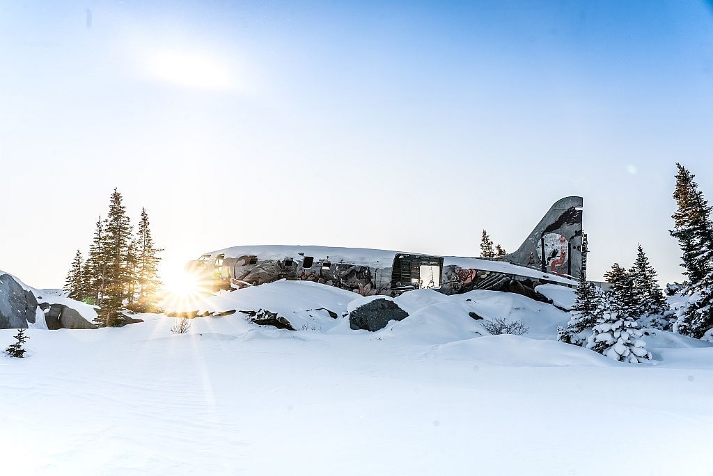 Sun sets behind snowy plane wreckage on a snow-covered mountain, surrounded by evergreen trees.