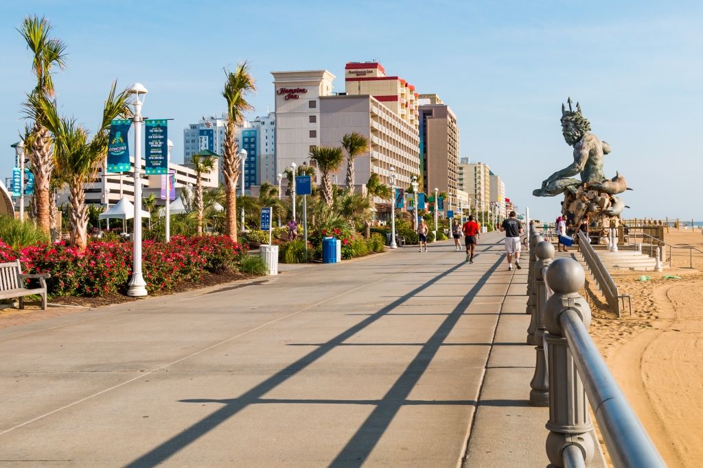 Beach boardwalk in Virginia, palm trees, hotels, and a statue. People walk and bike on a sunny day.