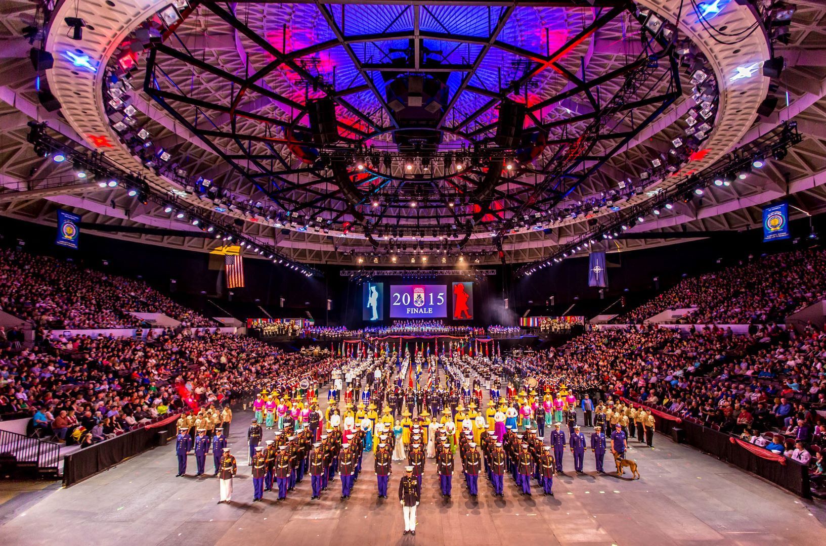 Large crowd watches a marching band performance in an arena, lit with colorful lights.