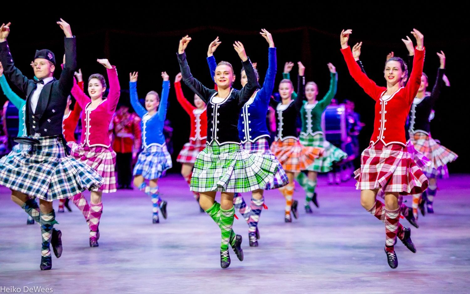 Scottish Highland dancers in colorful tartan skirts and jackets, arms raised, performing on stage.