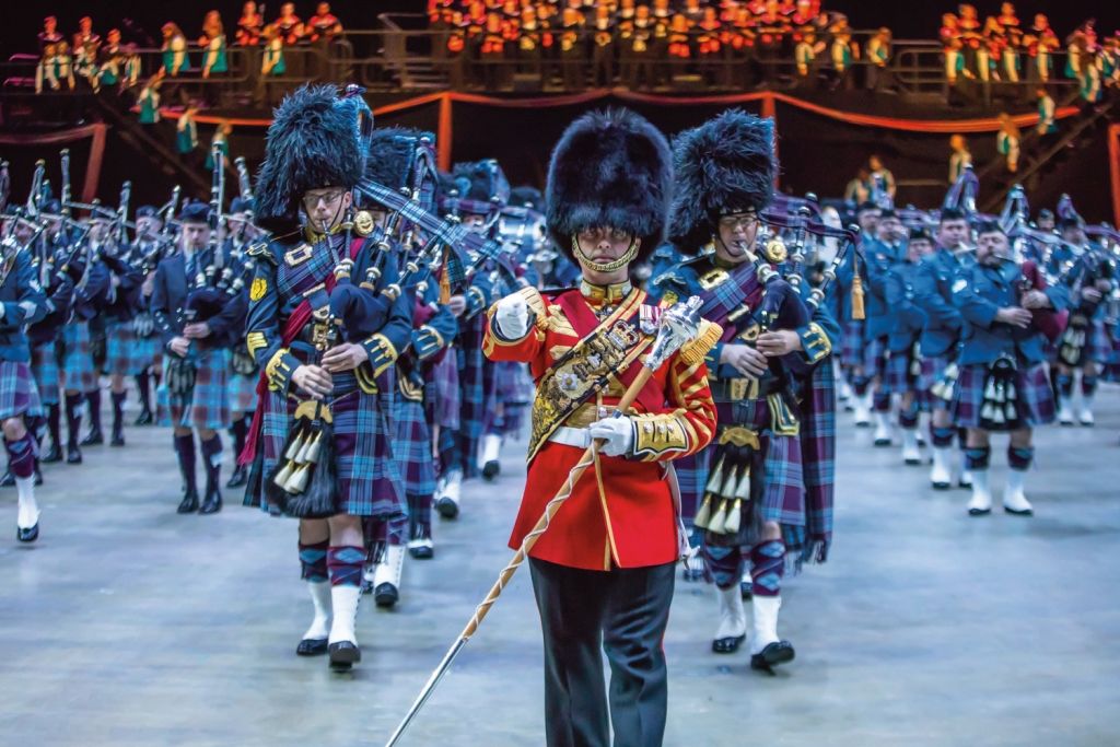 Scottish musicians in kilts and hats performing with a conductor in red, on a stage.