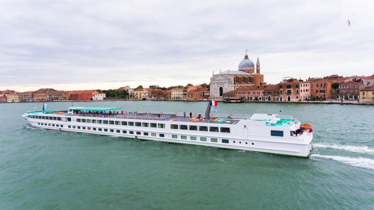 A white cruise ship sailing through a canal in Venice, Italy, with buildings in the background.