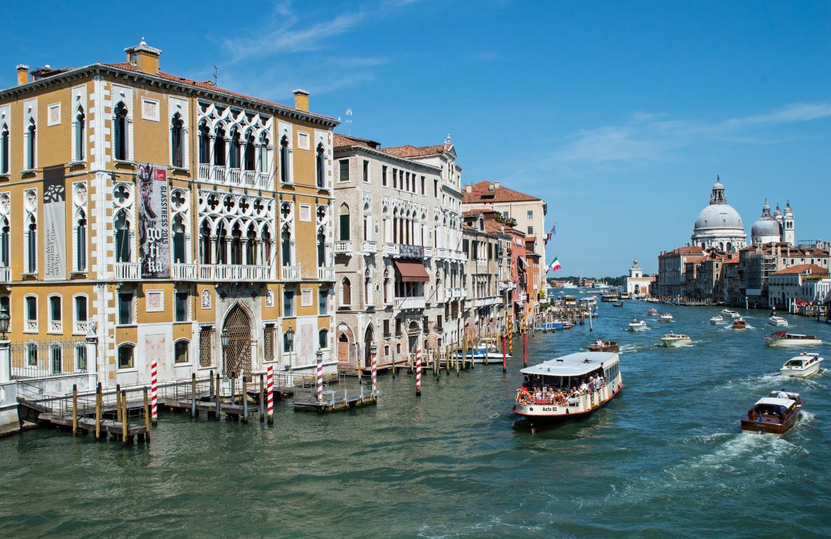 Venice canal with ornate buildings, boats, and a church under a blue sky.