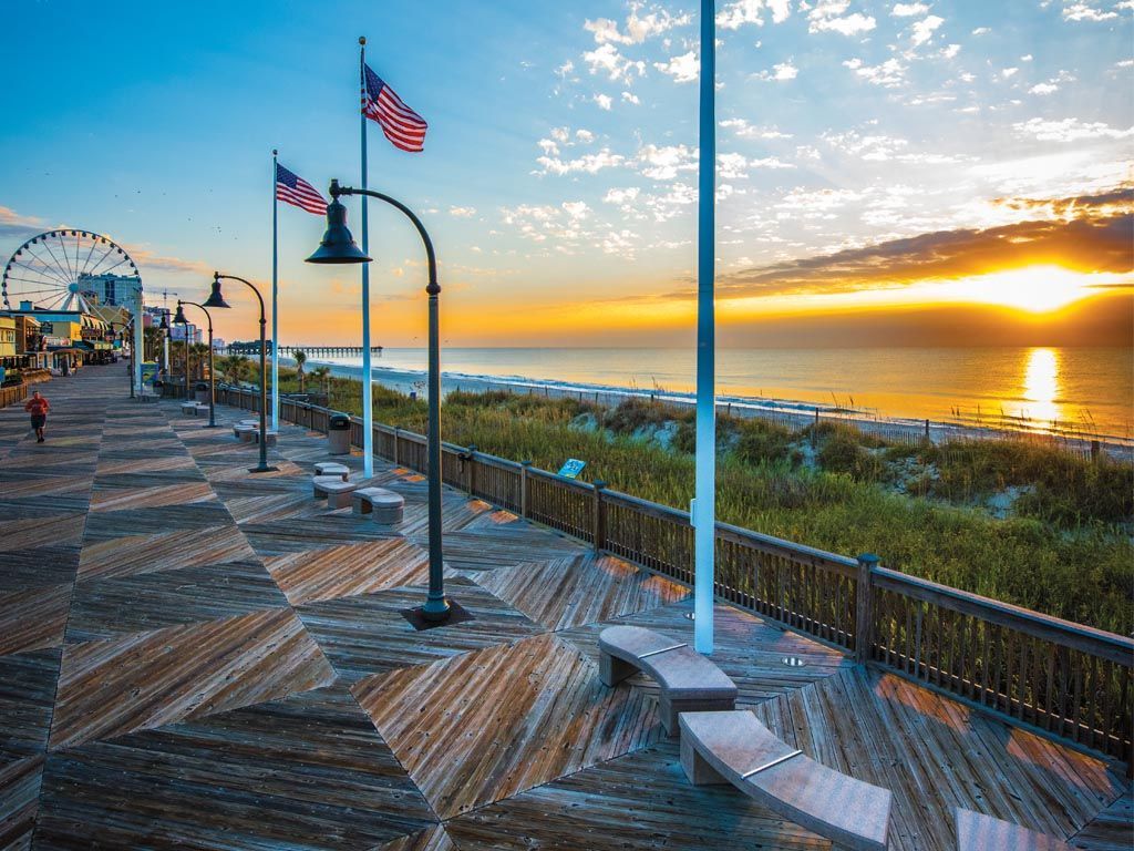 Wooden boardwalk with ocean view at sunset; American flags, streetlights, and Ferris wheel.