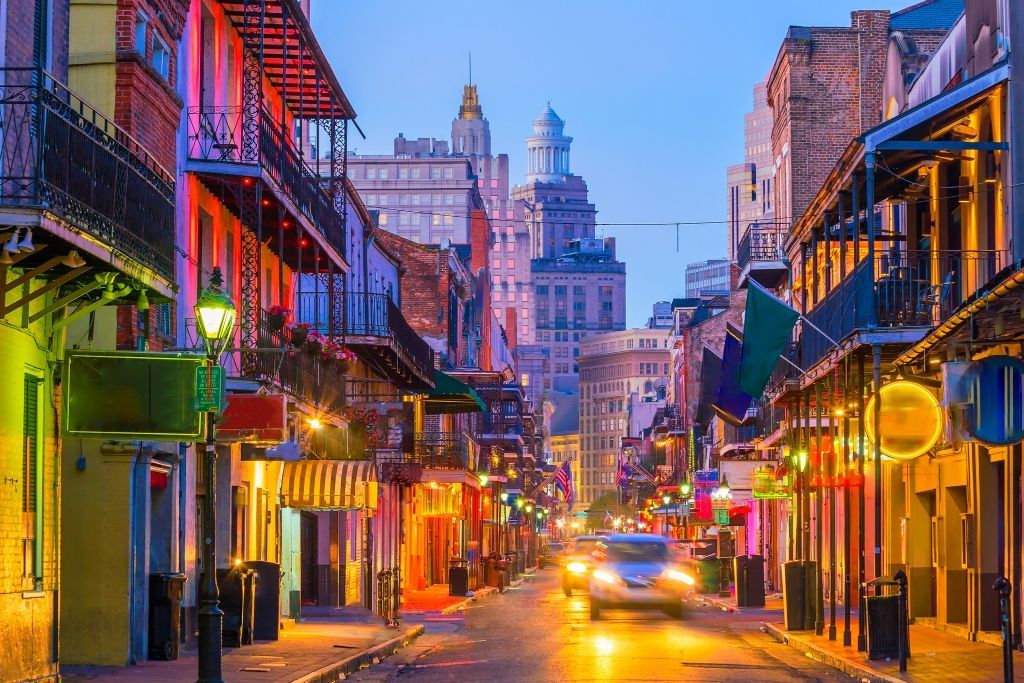 Colorful, lit-up street in New Orleans at dusk, with buildings and a car.