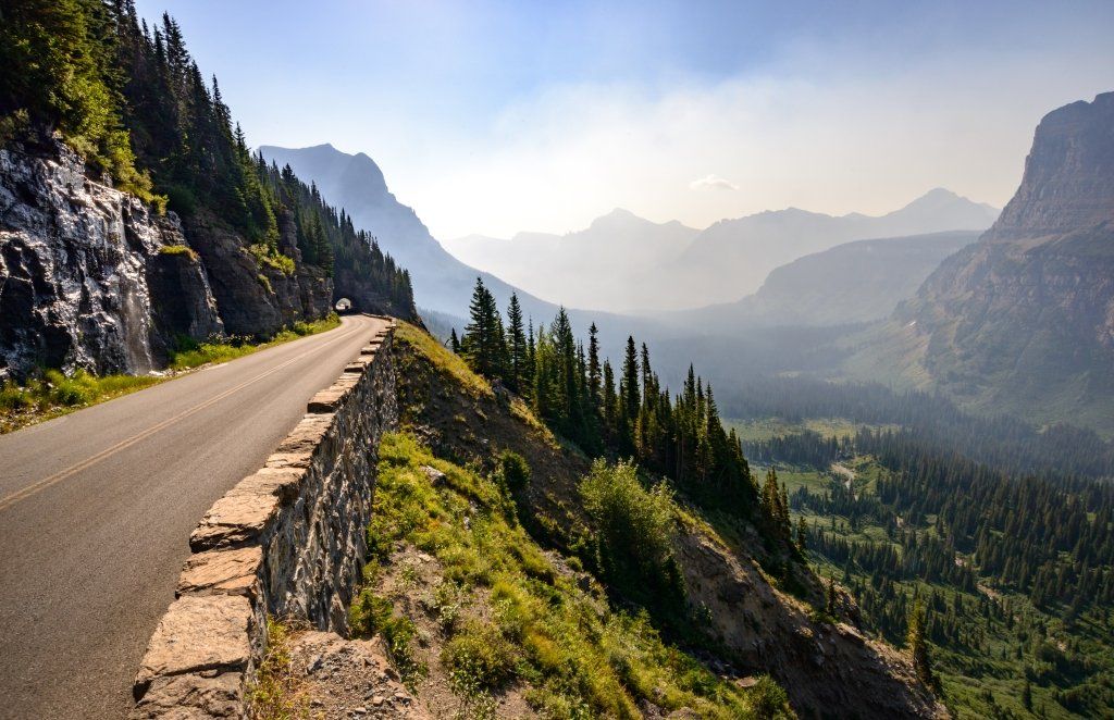 Mountain road winding through a scenic valley, flanked by rock walls and evergreen trees under a hazy sky.