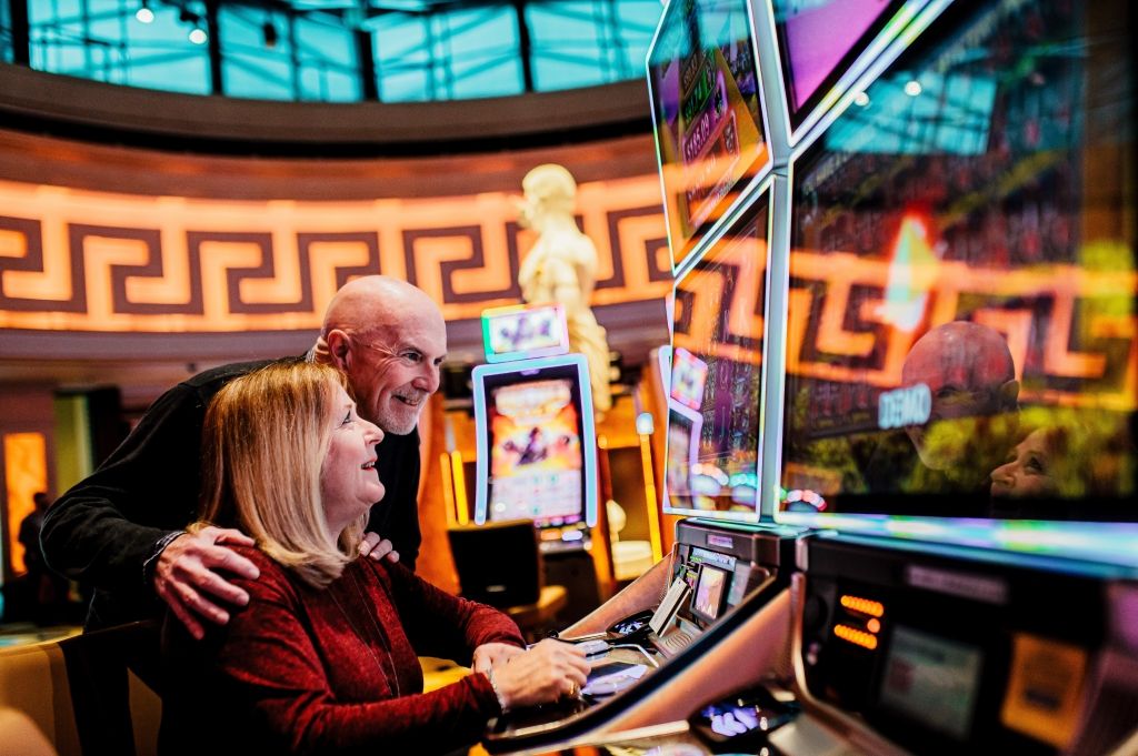 Older couple smiling at a slot machine in a casino, gold and red tones.