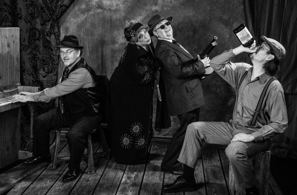 Four people in vintage clothing sit and stand indoors, drinking and playing music around a table.