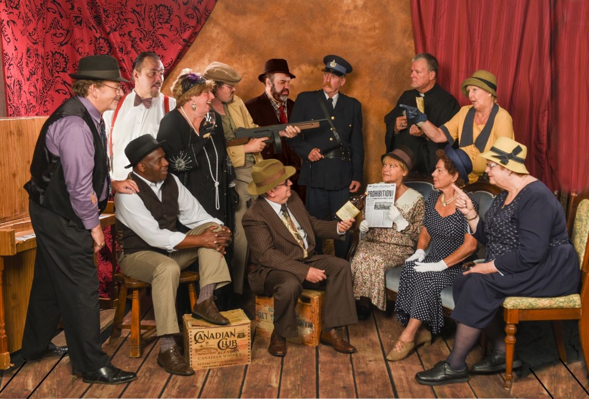 Group of people in vintage attire; some holding props like guns.  Indoor setting, likely a themed event.