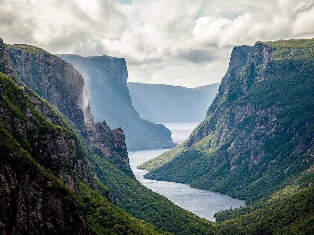 Dramatic mountain gorge with a lake winding through, green slopes, grey cliffs, and partly cloudy sky.