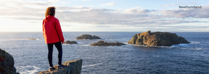 Person in a red jacket stands on a rocky cliff overlooking the ocean and small islands at sunset.