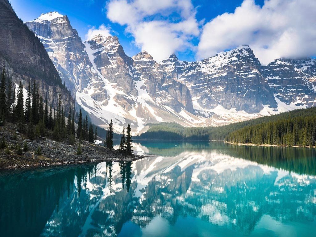 Snow-capped mountains reflected in a turquoise lake, surrounded by trees. Bright sky with clouds.