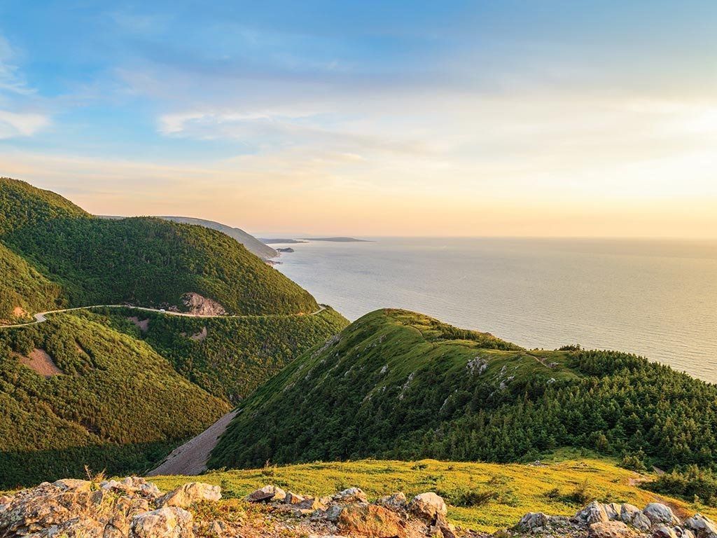 Green hills meet the ocean under a blue and gold sky; road curves along the coast.