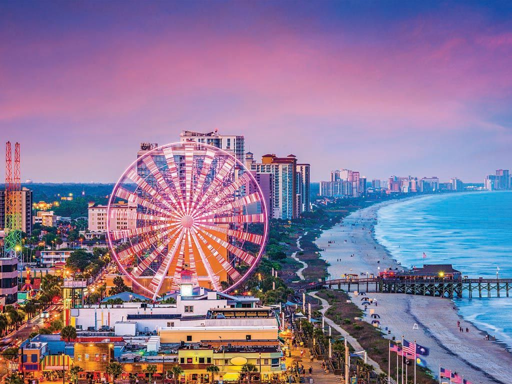 Ferris wheel lit up on Myrtle Beach, South Carolina, coastline at dusk with ocean and city buildings.