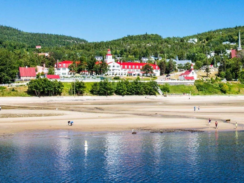 Beachfront view: white buildings with red roofs, green hills in the background, and blue water.