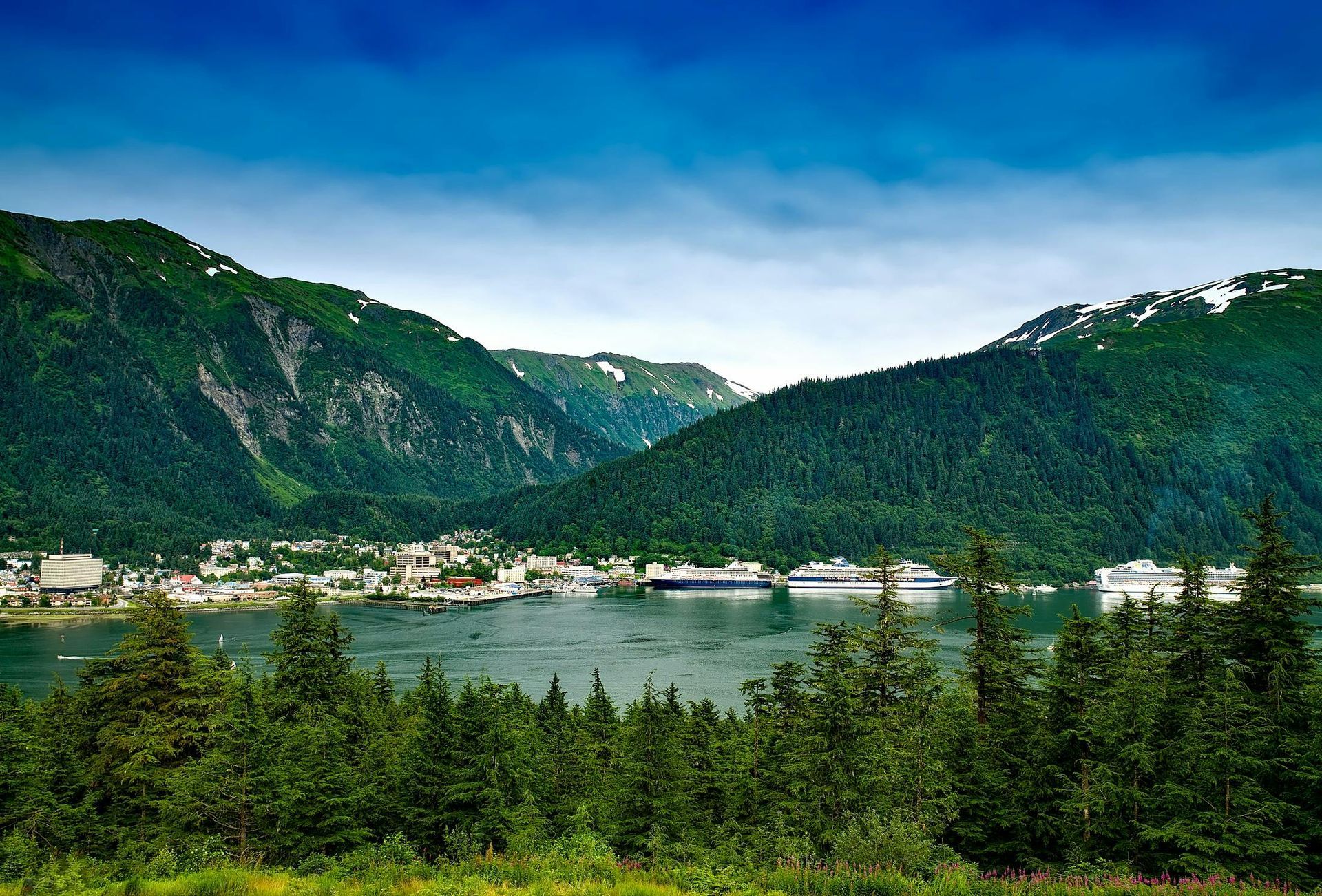 Coastal town nestled between lush green mountains and the blue ocean. Cruise ships, green trees, and cloudy blue sky.