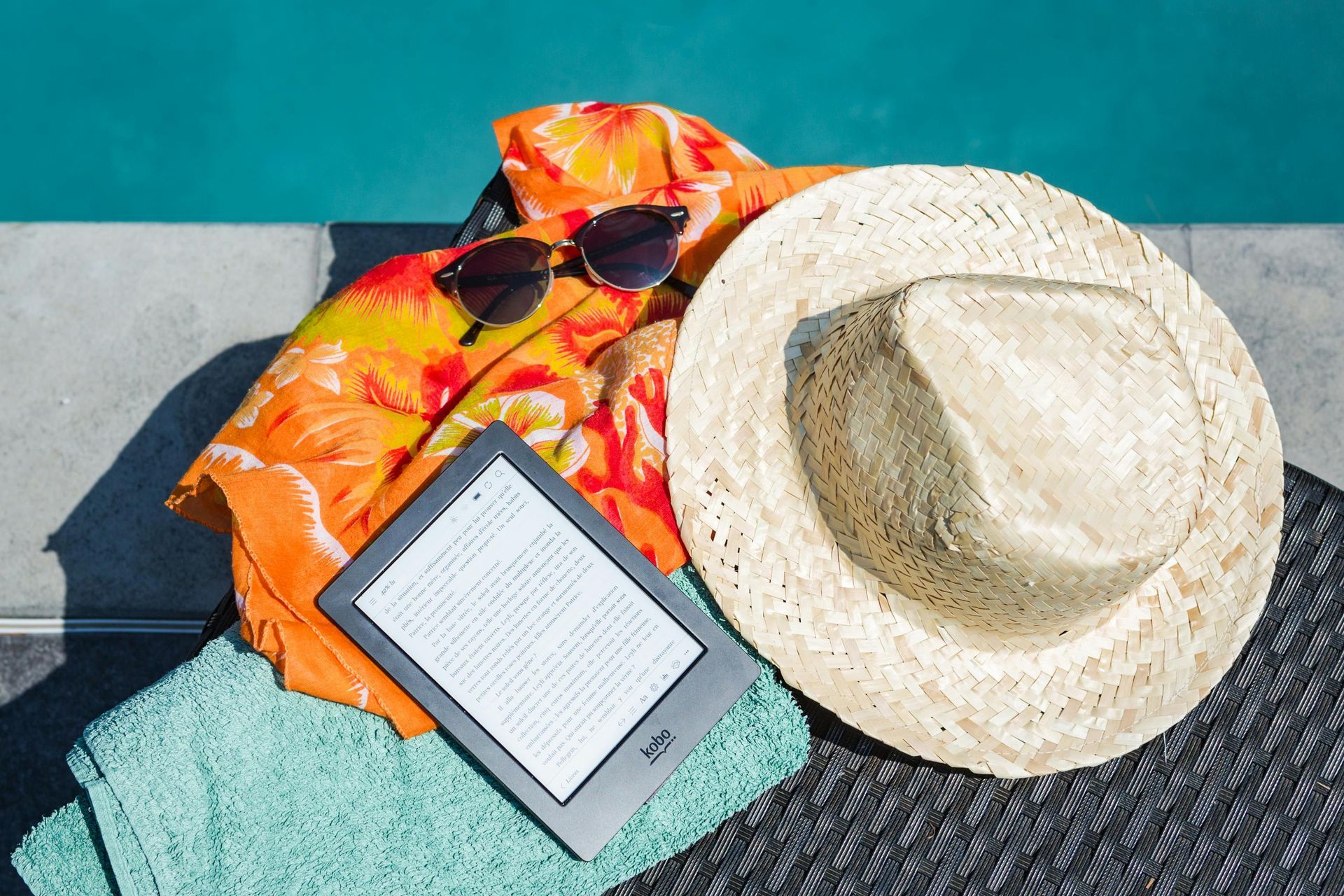 Sunhat, sunglasses, beach towel, and e-reader on a woven mat by a turquoise wall