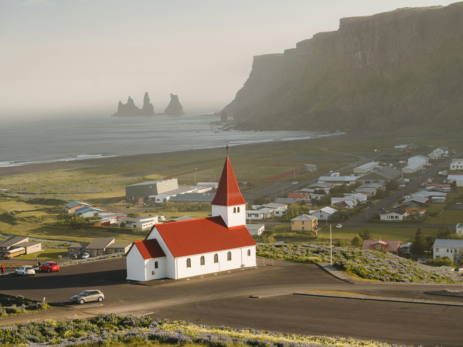 White church with red roof in a coastal town, with mountains and the ocean in the background.