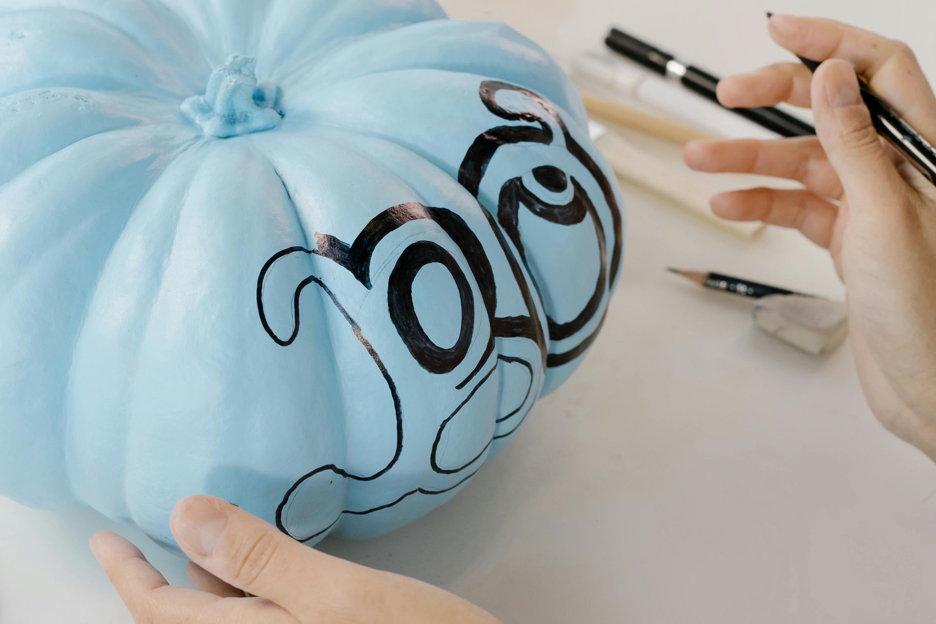 Person decorating a light blue pumpkin with black marker, on a white surface.