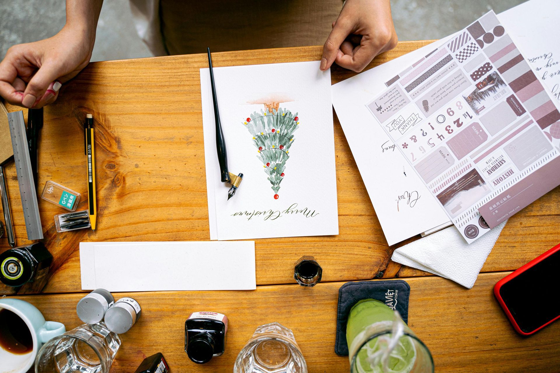 Person working on an art project at a wooden table, including watercolor art, pens, and various materials.