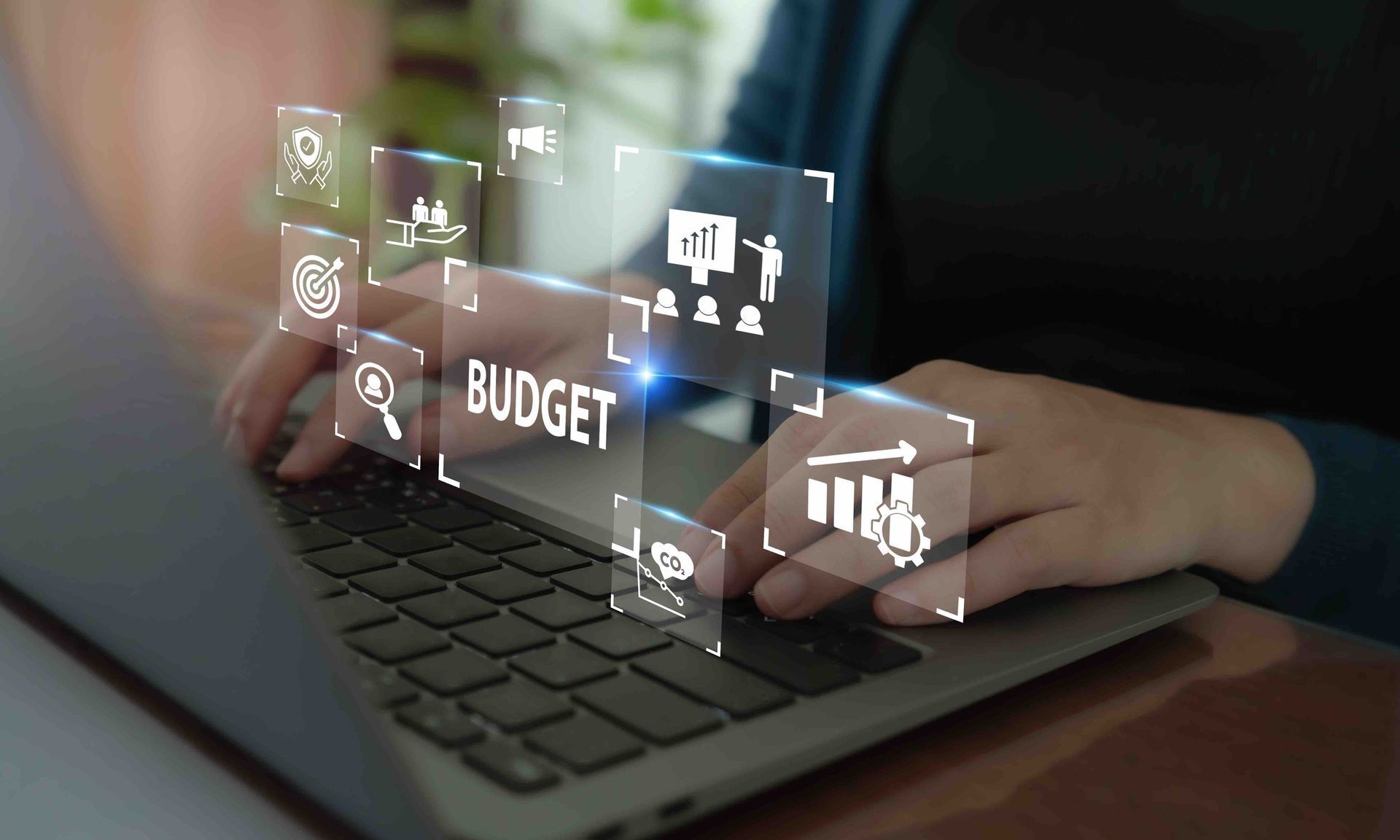 Hands typing on a laptop with a holographic budget and analytics interface hovering above the keyboard.