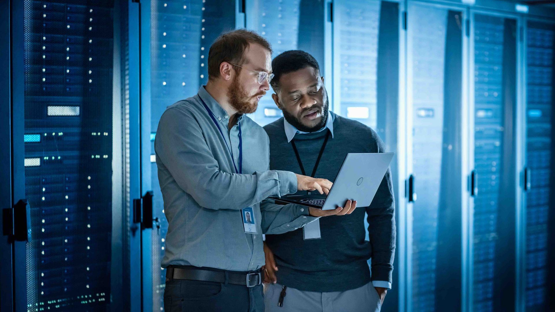 Two people in a server room reviewing a laptop beside blue-lit data racks