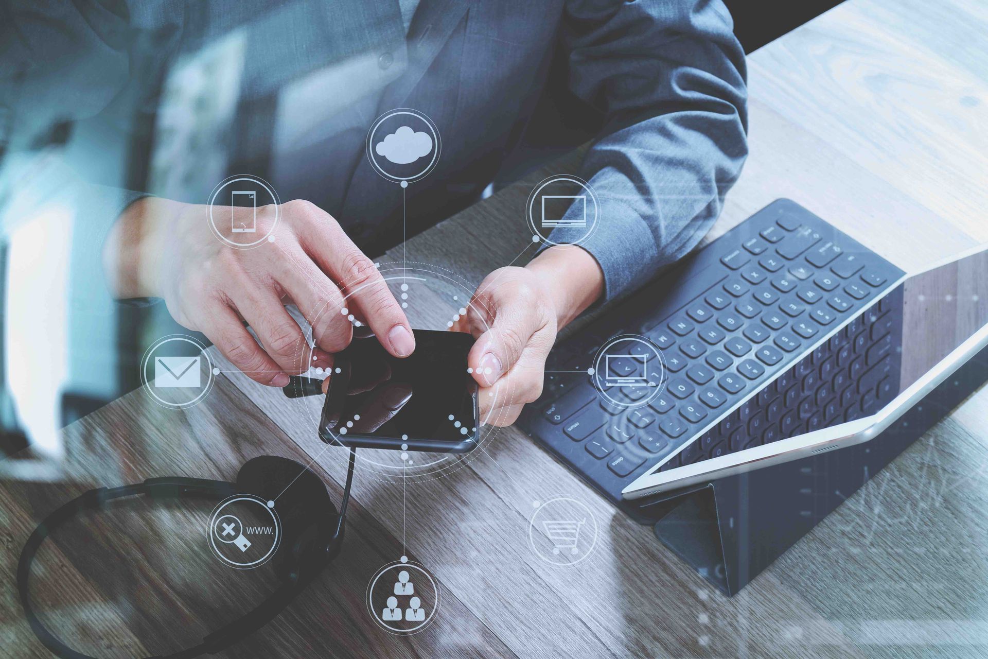 Person using a smartphone beside a laptop and keyboard on a desk with digital network icons.