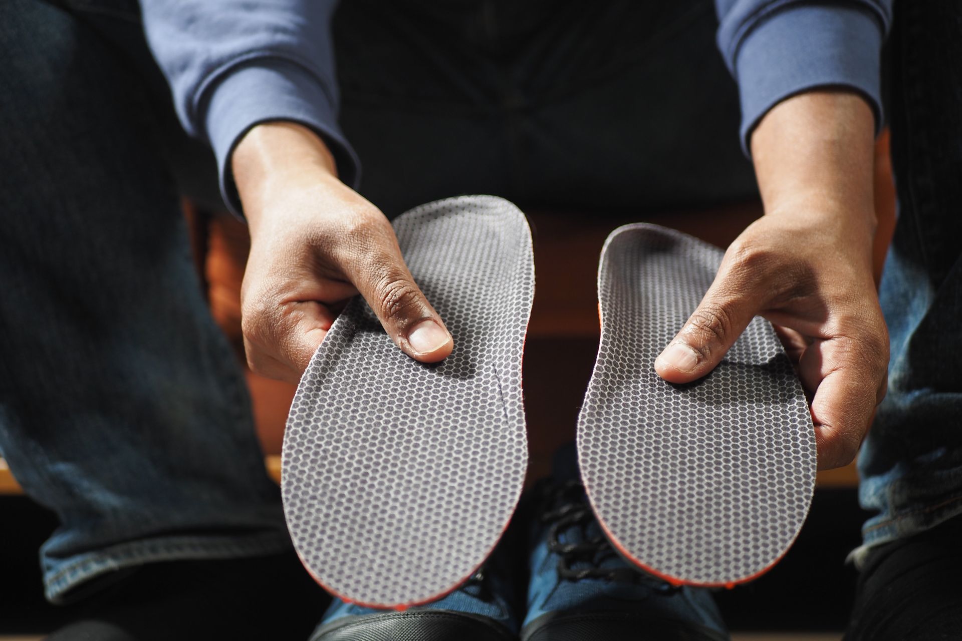 Hands holding a pair of gray shoe insoles with orange edges over jeans and sneakers.