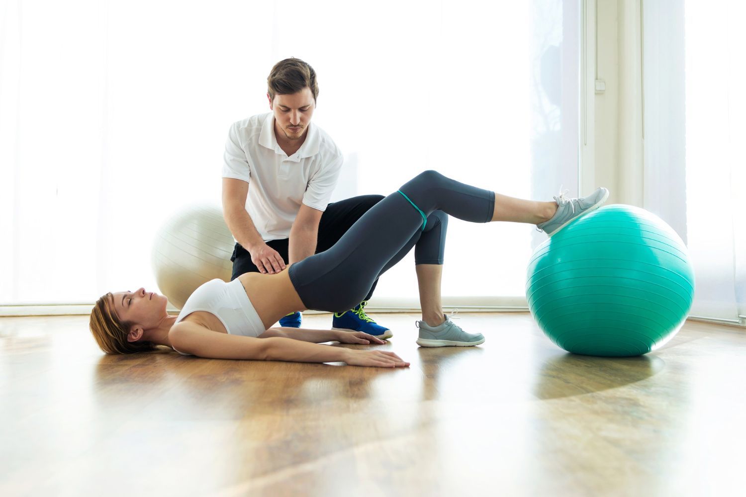 A physical therapist guides a person performing a glute bridge exercise with their feet resting on a large exercise ball.