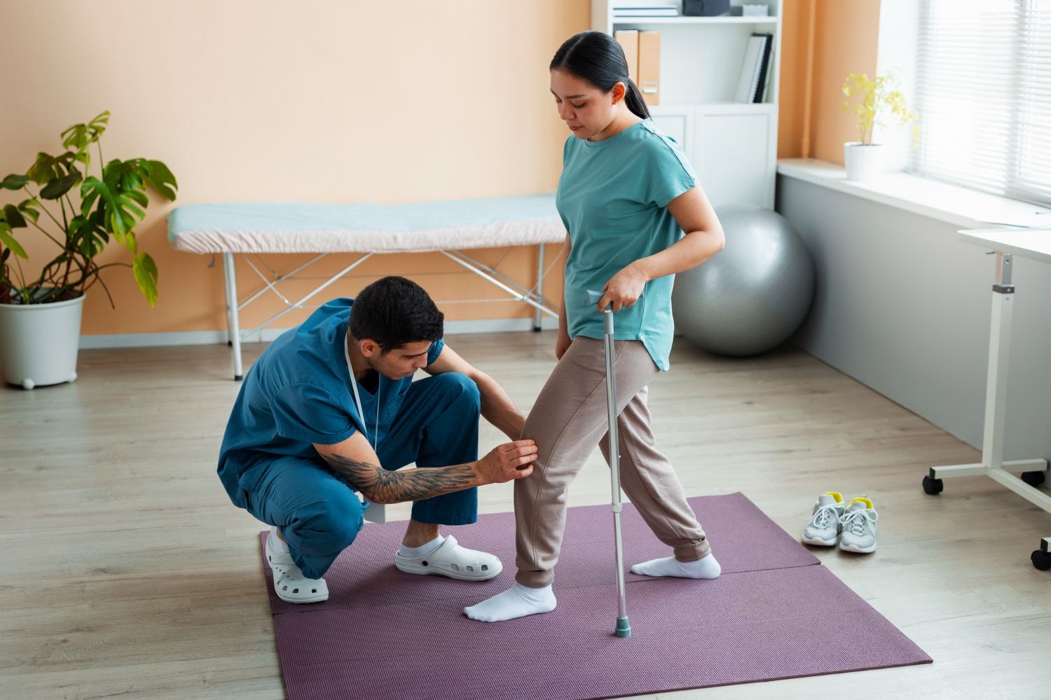 A physical therapist in blue scrubs examines a patient's knee as they stand on a mat using a cane in a clinic.