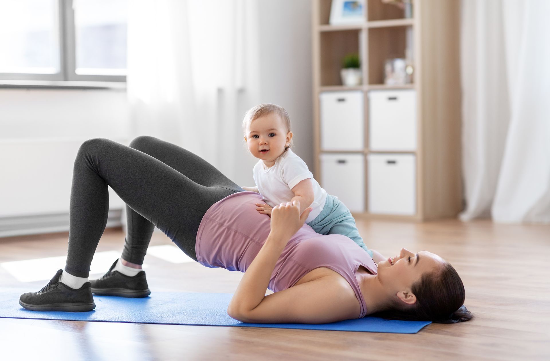 Woman doing a bridge pose on a yoga mat with a baby on her stomach in a bright room