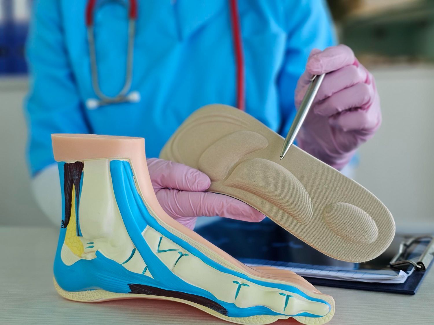 A medical professional in a blue top and pink gloves points to a textured orthopedic shoe insole next to a foot model.