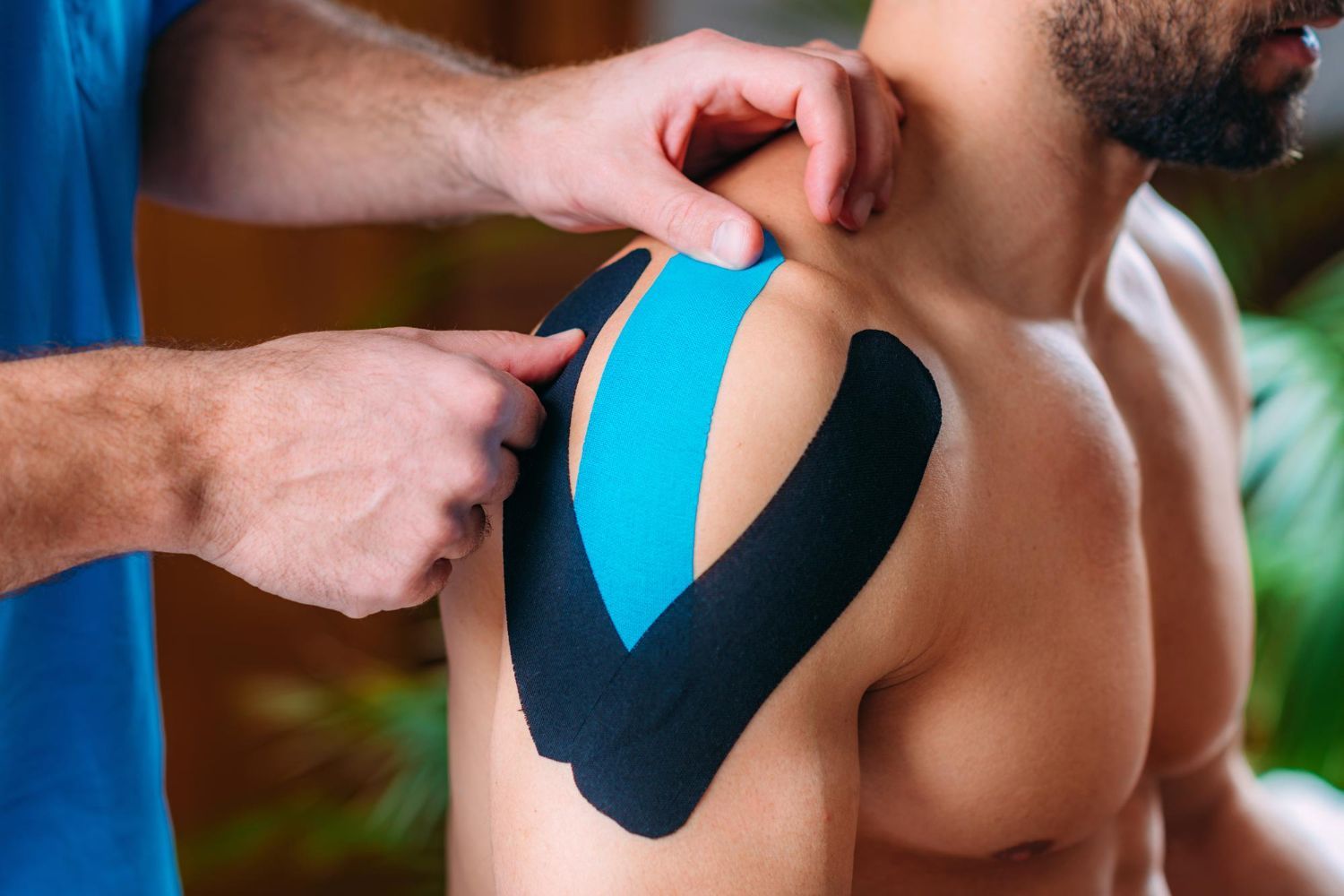 A professional applies blue and black kinesiology tape to a person's shoulder for physical therapy support.