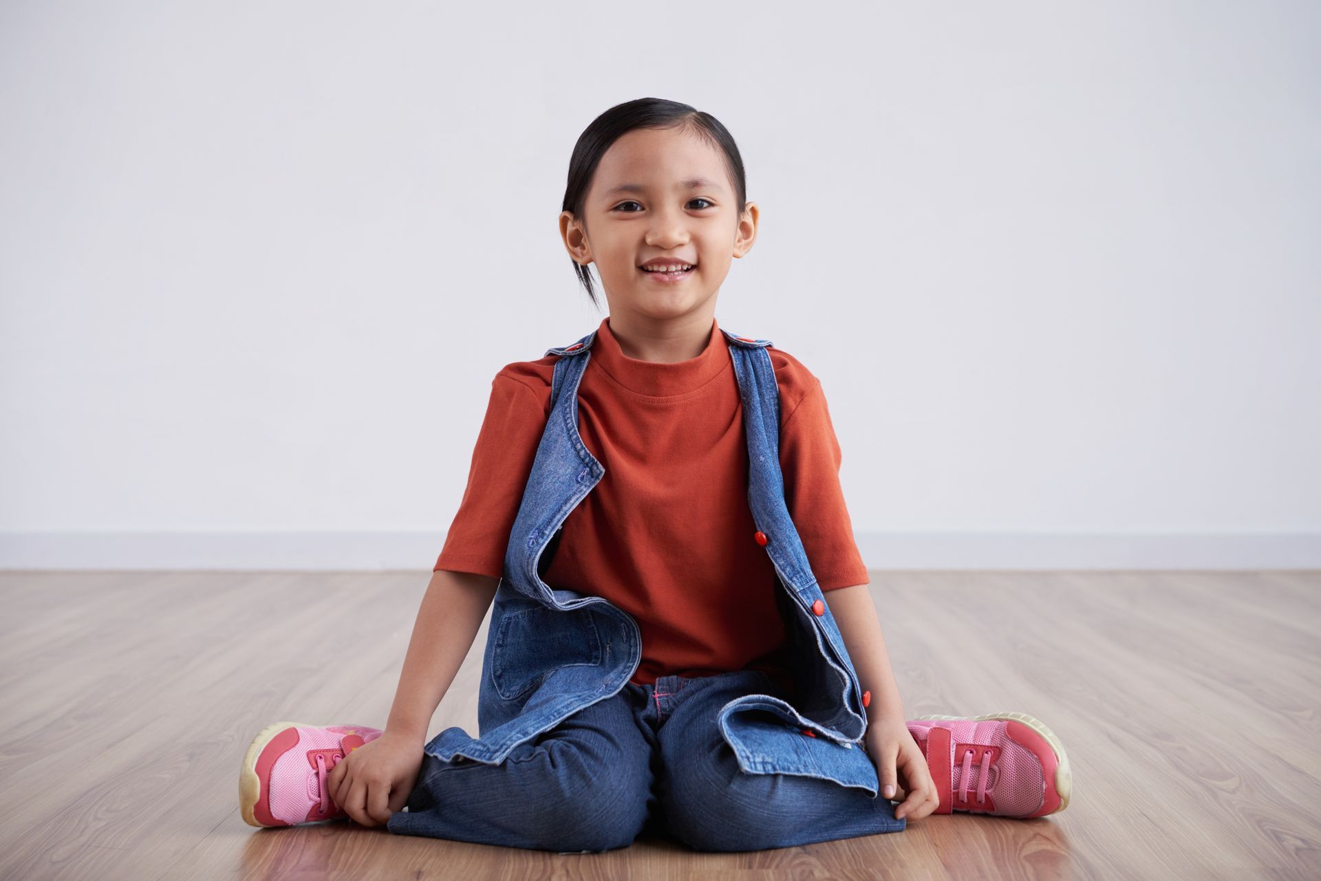 Smiling child sitting on the floor with a backpack, red shirt, jeans, and pink shoes in a bright room