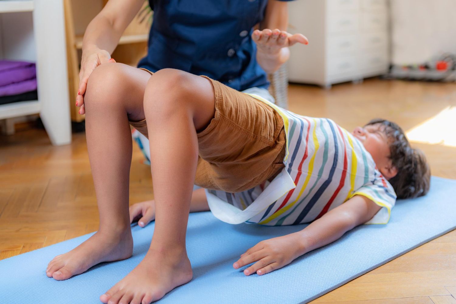A therapist guides a child performing a bridge exercise on a mat in a room.