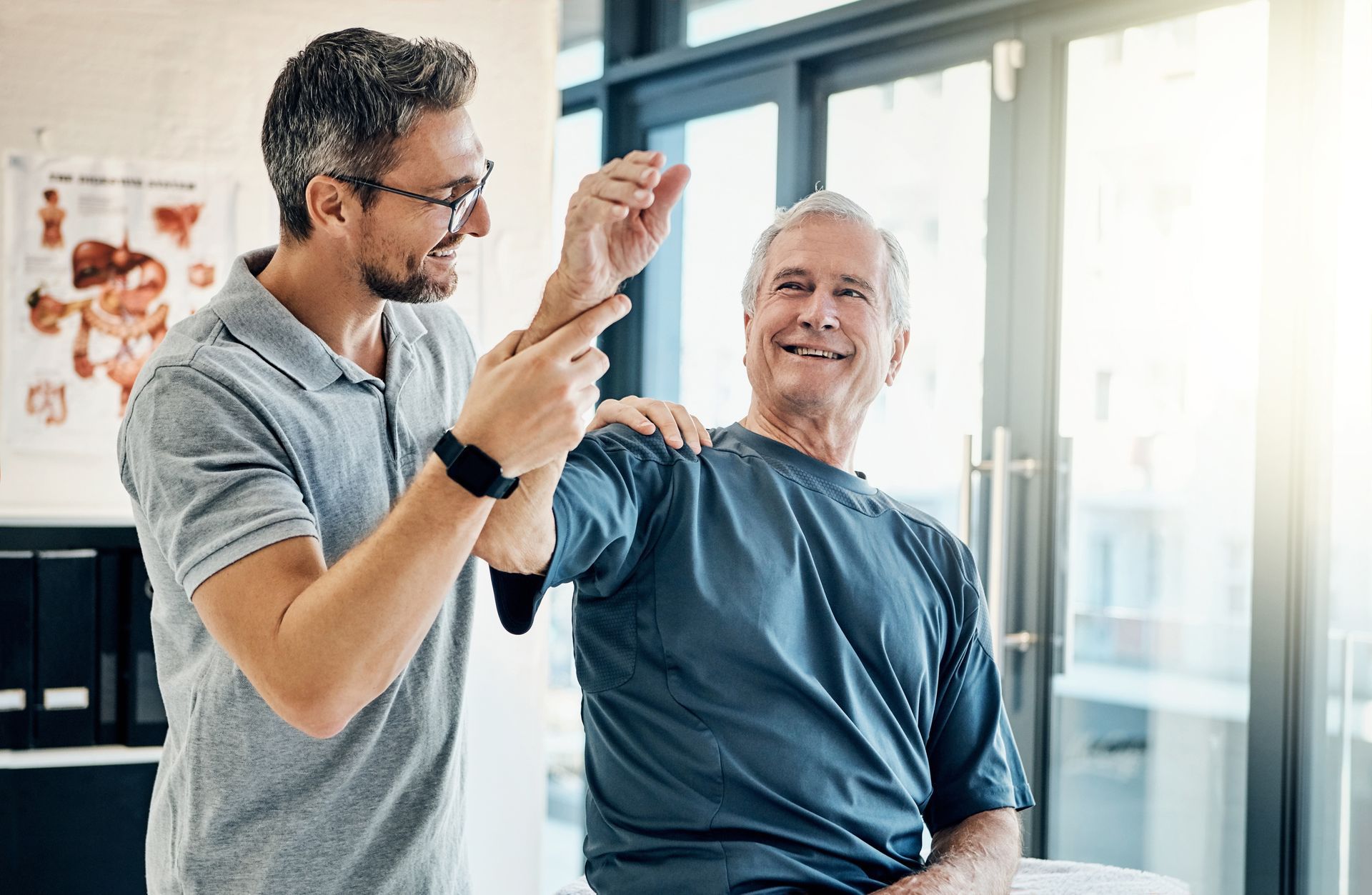 Two men in a bright clinic, one helping the other with an arm exercise and smiling