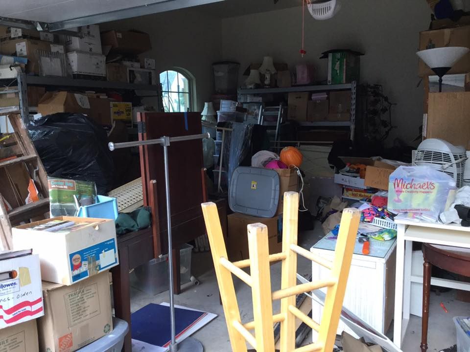 Messy garage filled with boxes, furniture, and clutter. Shelves are overflowing; a wooden stool sits overturned.