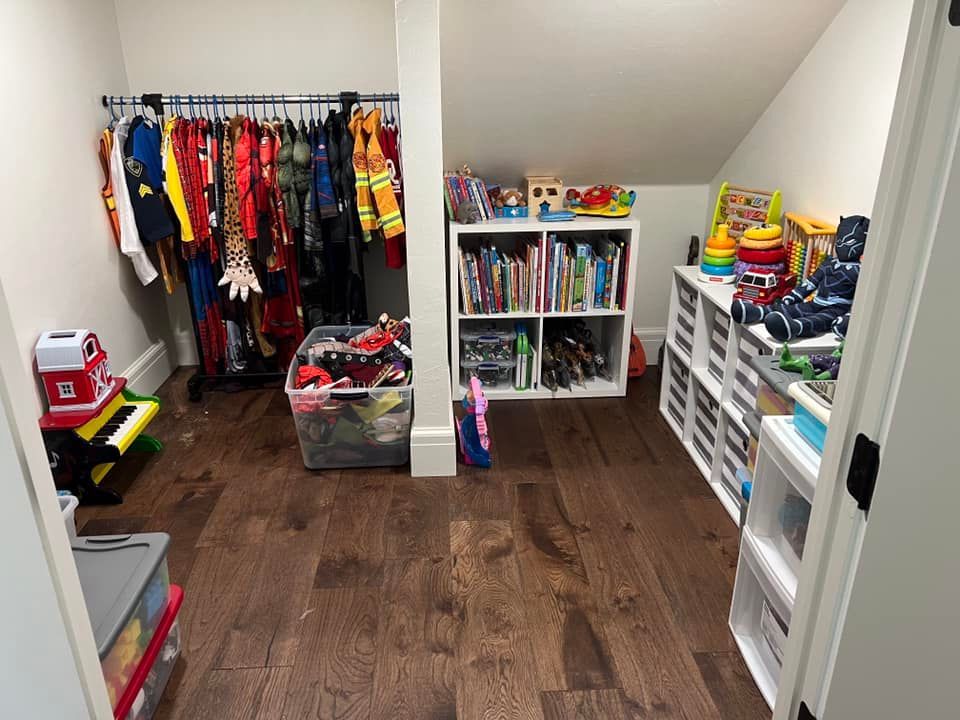 Small playroom with costumes hanging, bookshelves, and toy storage. Dark wooden floor.