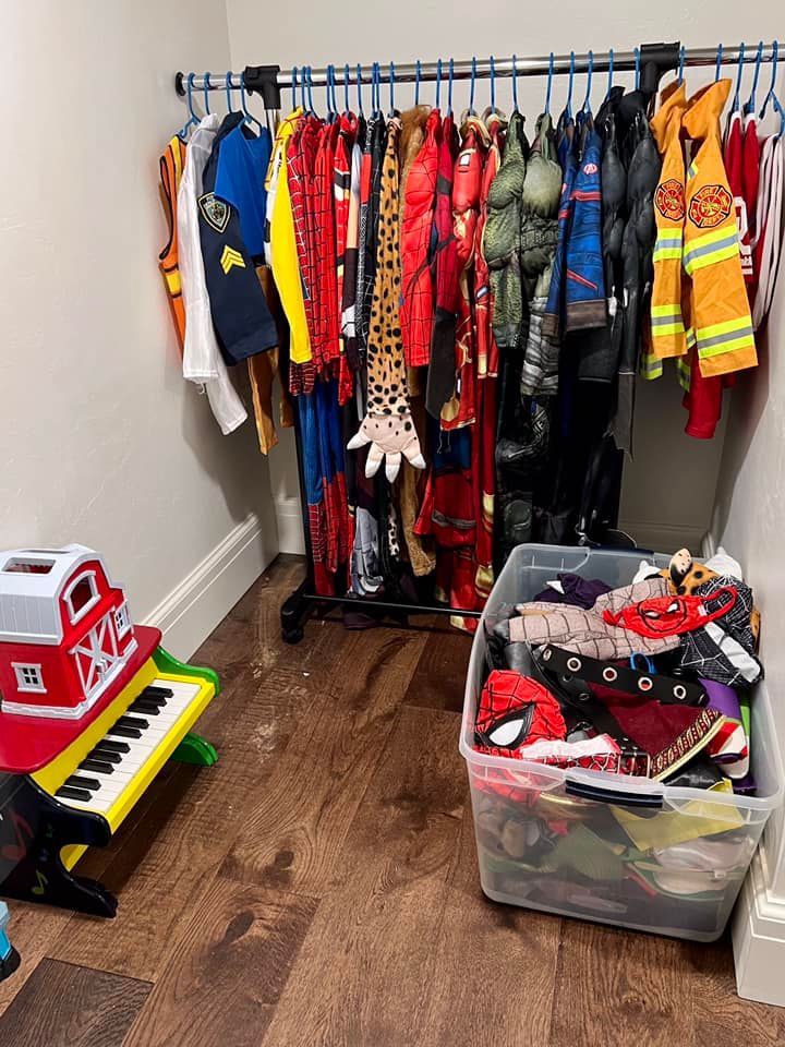 A child's costume closet. Costumes hang on a rack. A plastic bin of costumes sits below. A toy piano is on the left.
