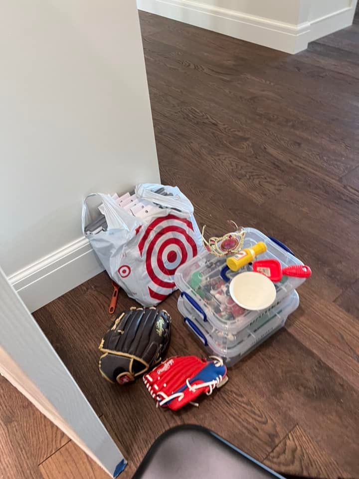 A Target bag, baseball gloves, and a plastic bin with toys sit on a wooden floor near a wall.