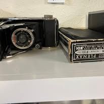 A vintage black camera with a lens and black film box on a white surface.